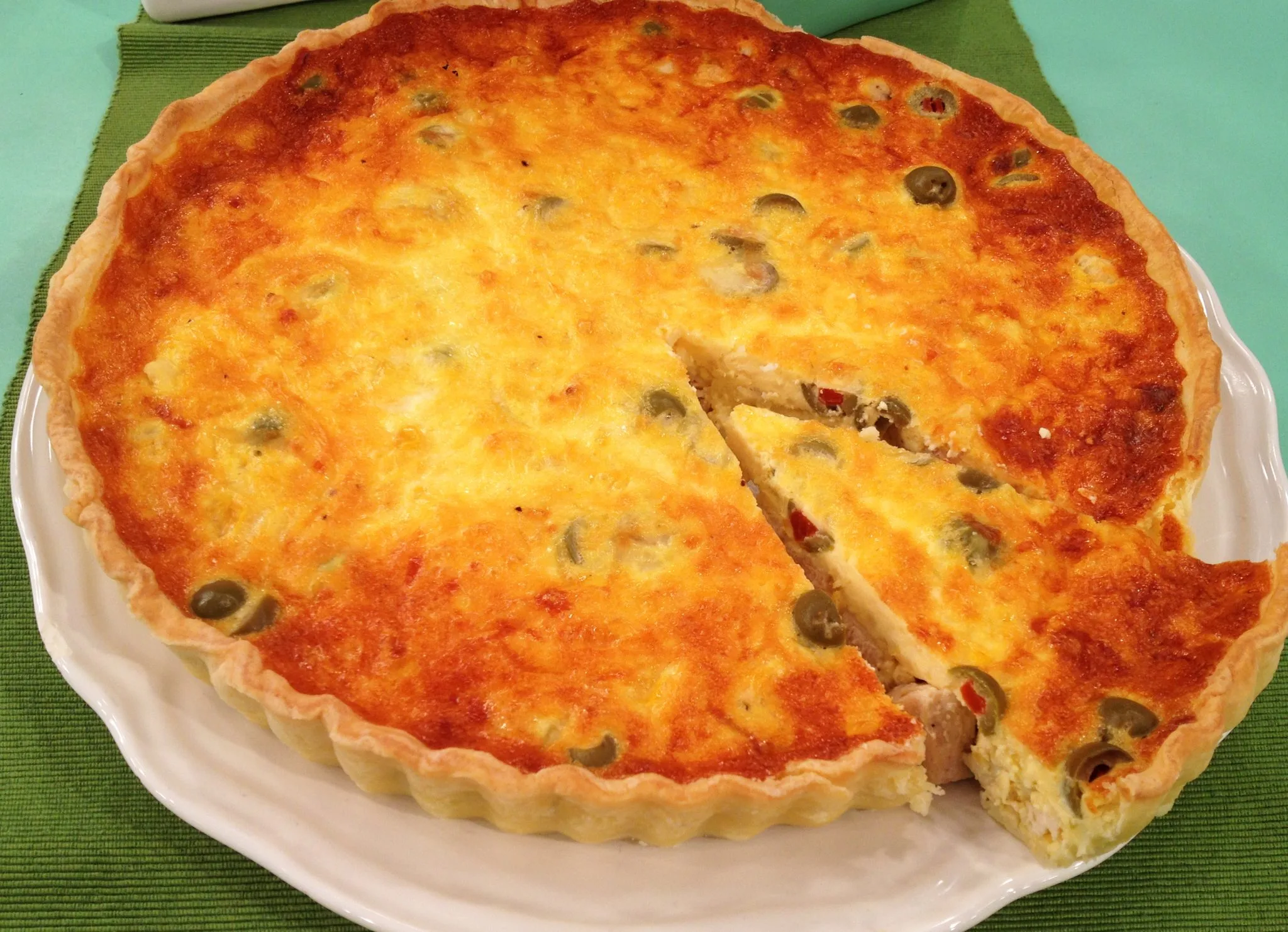 um quiche redondo em um prato redondo branco sobre uma tolha verde bandeira e um fundo azul claro