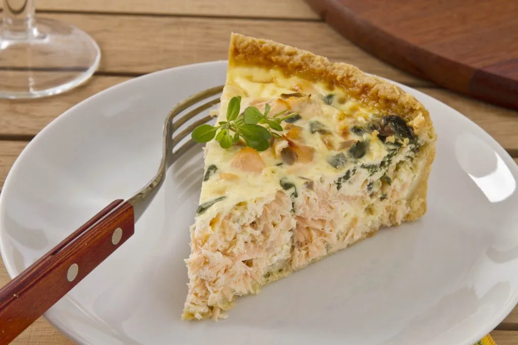 uma fatia de quiche de frango em um prato branco e um garfo de cabo de madeira, uma taça ao lado sobre uma mesa de madeira.