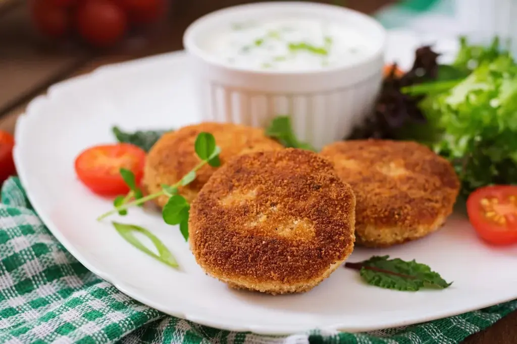 um prato com três bolinhos de couve-flor com temperos manjericão, hortelã rodelas de tomate, uma cumbuca com molho,sobre uma tolha xadrez verde e branca.