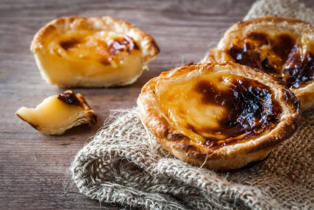 três Pasteis de Belém, um cortado, sobre um tecido rustico em uma mesa de madeira