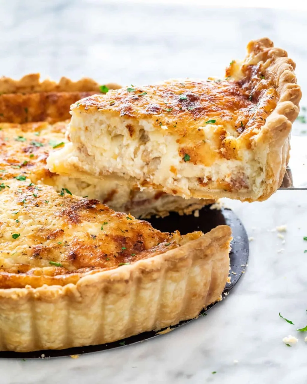 um quiche  lorraine em uma prato na cor preta, e uma fatia sendo  retirada com uma espátula, em um fundo marmorizado em cinza