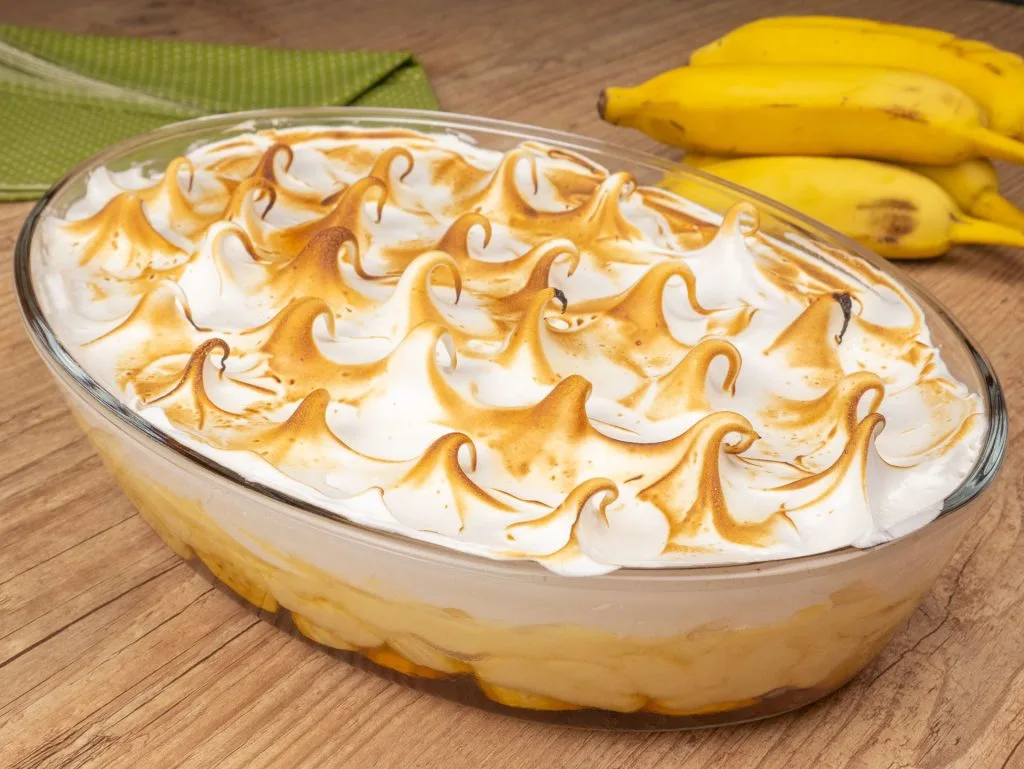 doce de banana com creme de leite em um refratário transparente, sobre uma mesa de madeira ao fundo algumas bananas