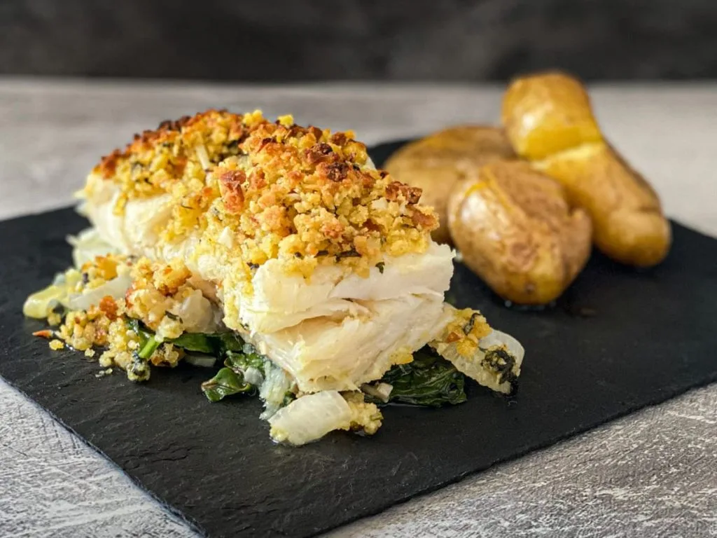 bacalhau com broa e espinafre, acompanhando batatas, sobre uma tabua de corte.