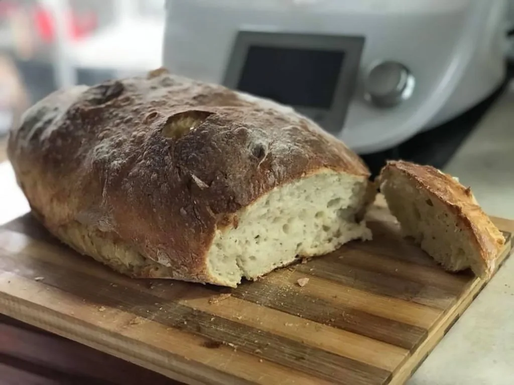 pão sobre uma tabua de corte, ja com uma  fatia retirada com uma bimby ao fundo