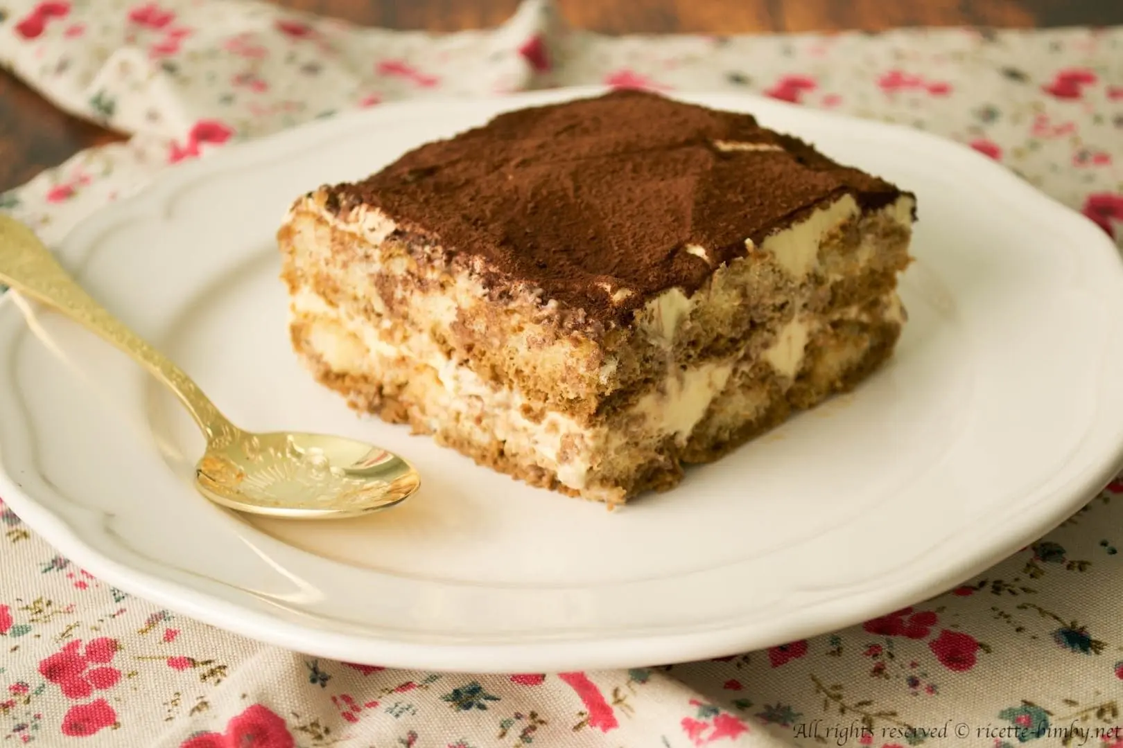 uma fatia de tiramisu dentro de um prato branco com uma colher ao lado