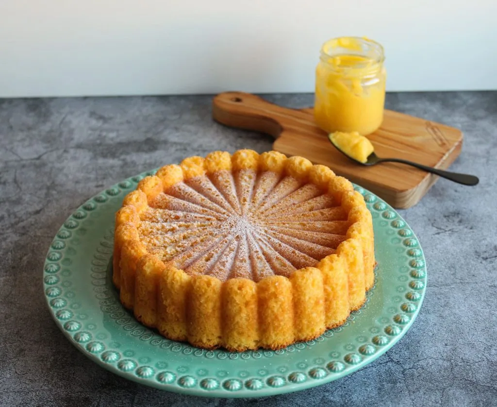 Bolo de limão em um prato na cor verde, ao fundo uma tabua de corte com um pote de doce e uma colher 