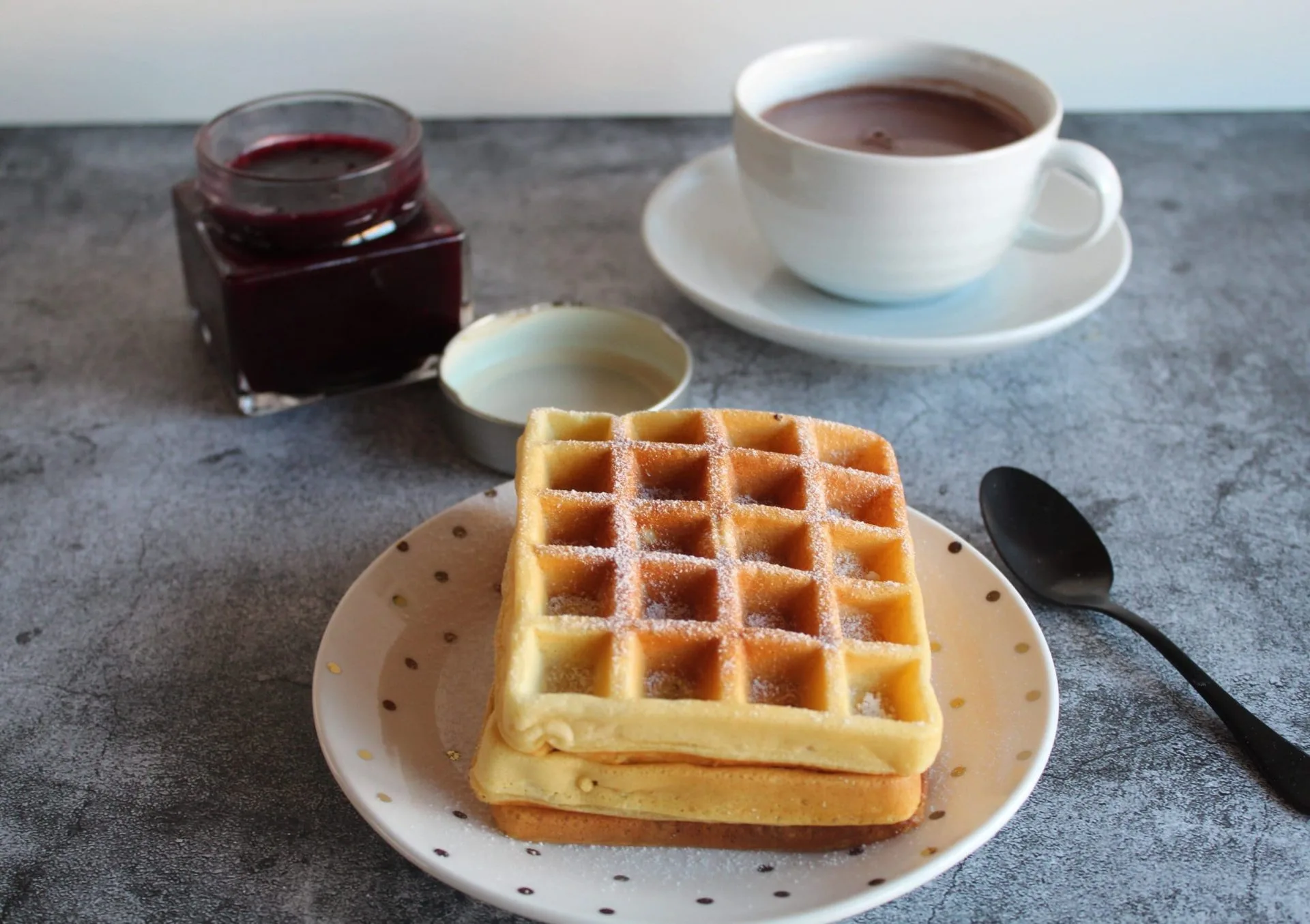 Waffles, sobre um prato branco ao lado talheres com um xicara com café ao fundo