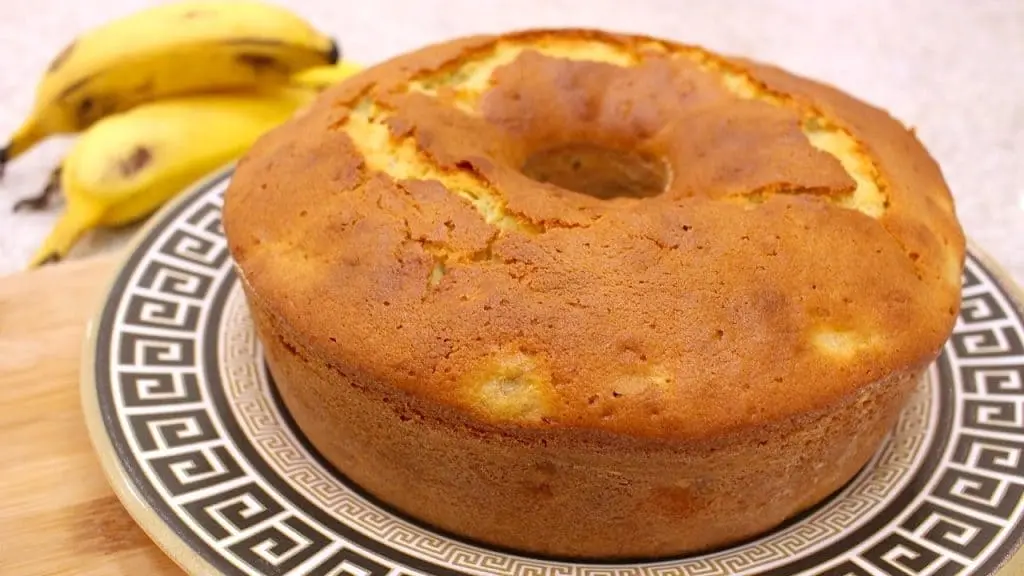 bolo de banana em um prato branco com detalhes em marrom com bananas ao fundo.