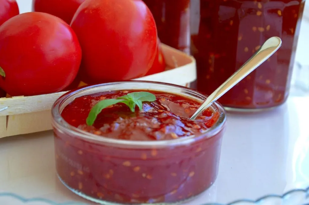 um pote com doce de tomate com uma colher dentro com alguns tomates ao fundo 