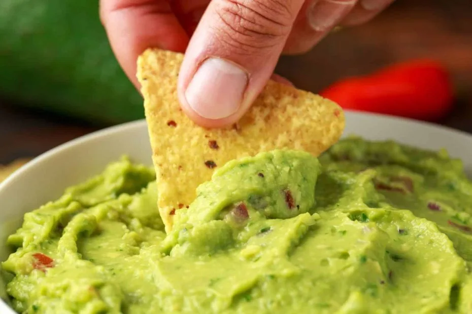 guacamole em uma travessa na cor branca, uma mão com uma tortilha retirando uma porção