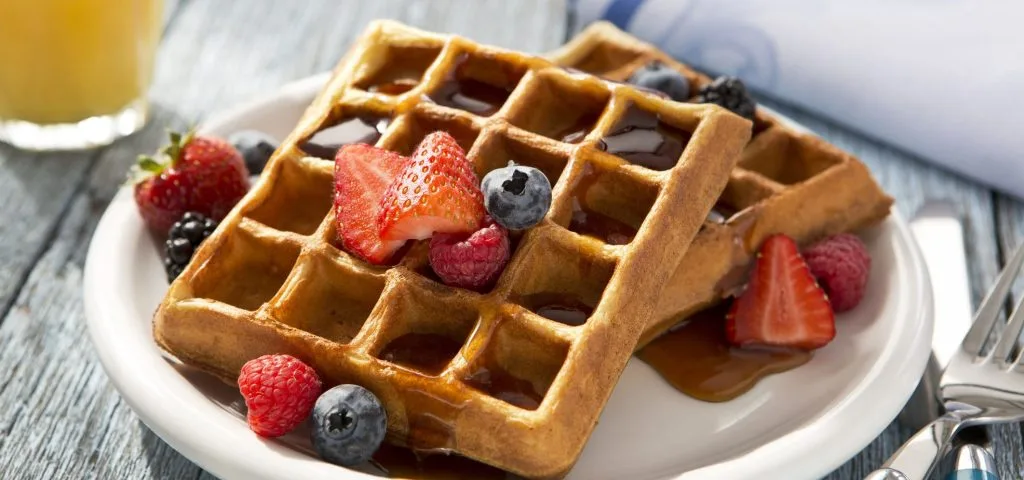 Waffles com calda e frutas, sobre um prato branco ao lado talheres com um copo com suco ao fundo