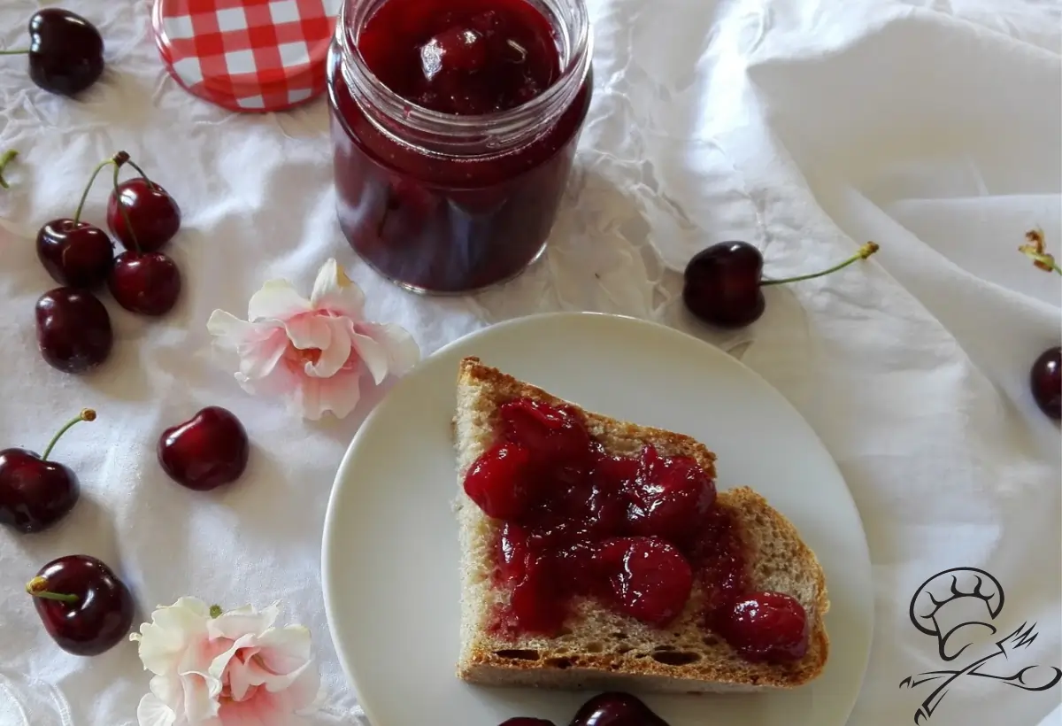 um vidro com doce de cereja ao lado um prato com uma fatia de pães com o doce com cerejas em torno